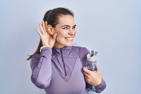 Photo for Beautiful woman wearing sportswear holding water bottle smiling with hand over ear listening an hearing to rumor or gossip. deafness concept. - Royalty Free Image