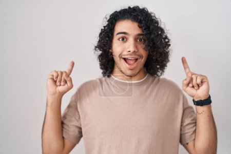 Téléchargez les photos : Homme hispanique aux cheveux bouclés debout sur fond blanc souriant étonné et surpris et pointant vers le haut avec les doigts et les bras levés. - en image libre de droit
