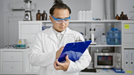 Photo for Serious young chinese man scientist, with a fashionable pigtail, diligently taking notes on a checklist in the meticulous world of lab research - Royalty Free Image
