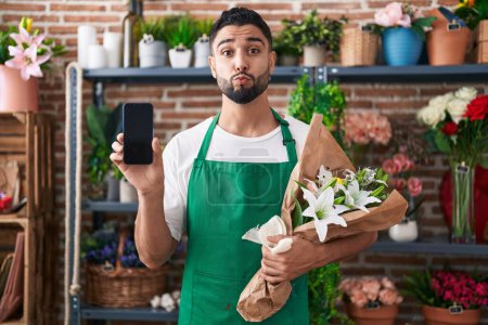 Photo for Hispanic young man working at florist shop showing smartphone screen looking at the camera blowing a kiss being lovely and sexy. love expression. - Royalty Free Image