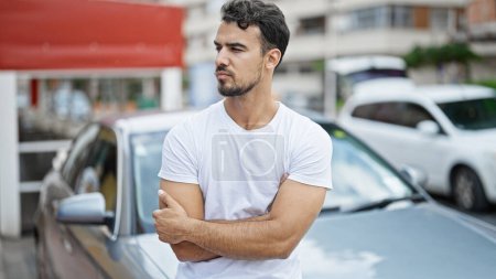 Téléchargez les photos : Jeune homme hispanique assis sur la voiture avec les bras croisés geste à la rue - en image libre de droit