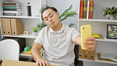 Téléchargez les photos : Beau jeune chinois, un employé de bureau sérieux mais détendu, capture un selfie professionnel avec son smartphone tout en travaillant à l'intérieur à son bureau exécutif dans un lieu de travail professionnel - en image libre de droit