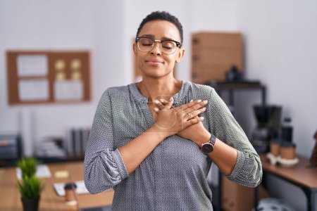 Foto de Mujer afroamericana trabajando en la oficina con gafas sonriendo con las manos en el pecho con los ojos cerrados y gesto agradecido en la cara. concepto de salud. - Imagen libre de derechos