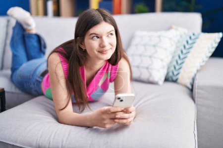 Téléchargez les photos : Jeune femme caucasienne utilisant smartphone couché sur canapé à la maison - en image libre de droit