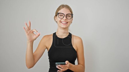 Foto de Mujer rubia joven usando un teléfono inteligente haciendo un gesto aceptable sobre un fondo blanco aislado - Imagen libre de derechos