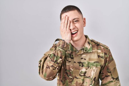 Téléchargez les photos : Jeune homme portant un uniforme de camouflage de l'armée couvrant un oeil avec la main, sourire confiant sur le visage et l'émotion surprise. - en image libre de droit