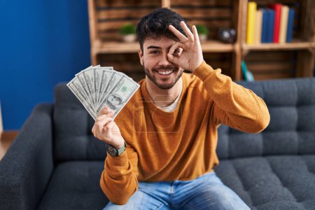 Foto de Hombre hispano con barba sosteniendo billetes de 100 dólares sonriendo feliz haciendo buen signo con la mano en el ojo mirando a través de los dedos - Imagen libre de derechos