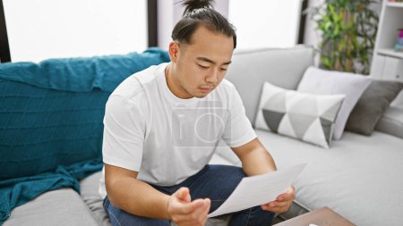 Photo for Upset young chinese man with smart expression intensely reading worrying document, sitting on sofa at home, indoors - Royalty Free Image
