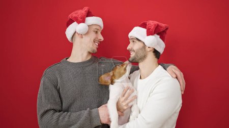 Photo for Two men couple wearing christmas hat hugging dog over isolated red background - Royalty Free Image