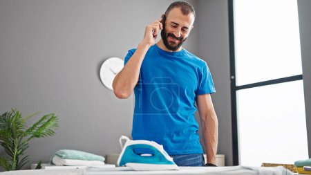 Photo for Young hispanic man talking on smartphone ironing clothes at laundry room - Royalty Free Image