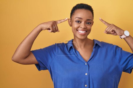 Foto de Mujer afroamericana de pie sobre fondo amarillo sonriendo señalando a la cabeza con ambas manos dedo, gran idea o pensamiento, buena memoria - Imagen libre de derechos