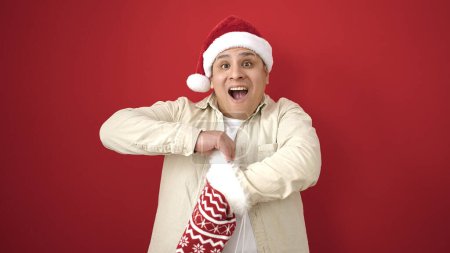 Téléchargez les photos : Jeune homme hispanique portant un chapeau de Noël regardant à l'intérieur de la chaussette sur fond rouge isolé - en image libre de droit