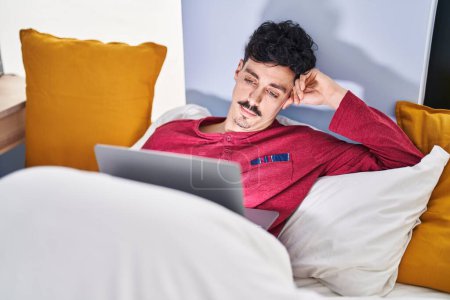 Téléchargez les photos : Jeune homme caucasien regardant un film assis sur le lit à la chambre - en image libre de droit