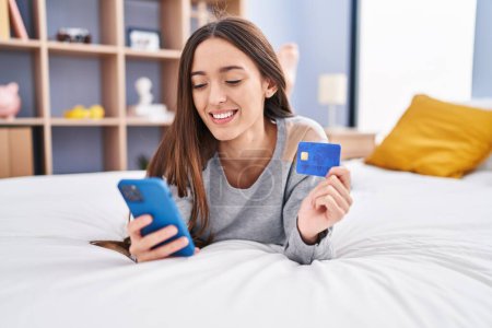 Téléchargez les photos : Jeune belle femme hispanique utilisant smartphone et carte de crédit couché sur le lit dans la chambre - en image libre de droit