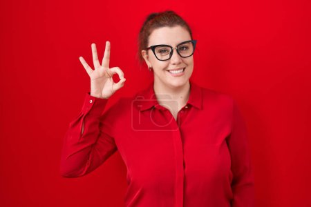 Foto de Mujer hispana joven con el pelo rojo de pie sobre fondo rojo sonriendo positiva haciendo signo de ok con la mano y los dedos. expresión exitosa. - Imagen libre de derechos