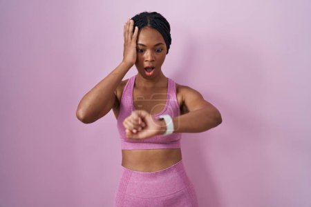 Téléchargez les photos : Femme afro-américaine avec des tresses portant des vêtements de sport sur fond rose en regardant l'heure de la montre inquiet, peur de se retrouver en retard - en image libre de droit