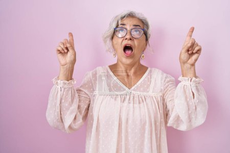 Foto de Middle age woman with grey hair standing over pink background amazed and surprised looking up and pointing with fingers and raised arms. - Imagen libre de derechos