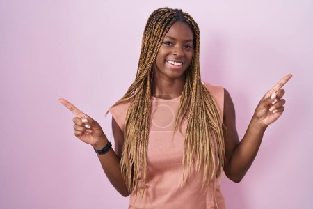 Téléchargez les photos : Femme afro-américaine avec les cheveux tressés debout sur fond rose souriant confiant pointant avec les doigts vers différentes directions. espace de copie pour la publicité - en image libre de droit
