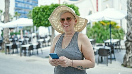 Foto de Turista mujer de pelo gris de mediana edad sonriendo confiada usando teléfono inteligente en la terraza de la cafetería - Imagen libre de derechos