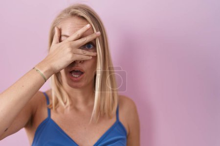 Foto de Mujer caucásica joven de pie sobre fondo rosa asomándose en shock cubriendo la cara y los ojos con la mano, mirando a través de los dedos con expresión avergonzada. - Imagen libre de derechos