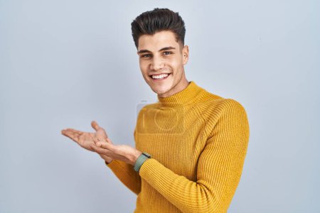 Foto de Joven hombre hispano de pie sobre fondo azul invitando a entrar sonriendo natural con la mano abierta - Imagen libre de derechos