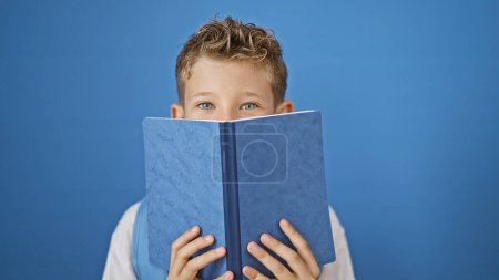 Photo for Adorable blonde boy student, mouth covered with book, standing isolated on a blue background, diving into learning with passion. - Royalty Free Image
