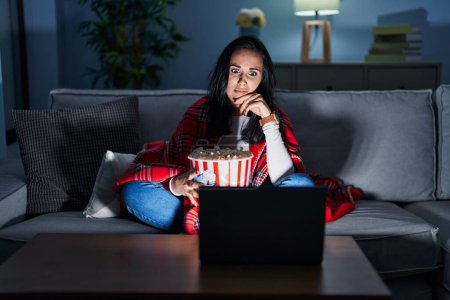 Téléchargez les photos : Femme hispanique manger du pop-corn regarder un film sur le canapé en pensant inquiet d'une question, préoccupé et nerveux avec la main sur le menton - en image libre de droit