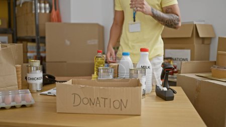 Téléchargez les photos : Jeune homme hispanique bénévole mettant des produits sur la boîte en carton de don au centre de charité - en image libre de droit