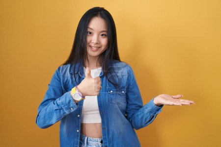 Téléchargez les photos : Jeune femme asiatique debout sur fond jaune montrant la main de la paume et faisant un geste correct avec les pouces levés, souriant heureux et joyeux - en image libre de droit