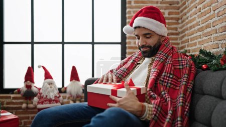 Photo for Young hispanic man wearing christmas hat unpacking gift at home - Royalty Free Image