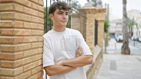 Téléchargez les photos : Jeune adolescent hispanique regardant vers le côté avec une expression sérieuse et les bras croisés geste à la rue - en image libre de droit