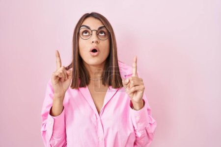 Téléchargez les photos : Jeune femme hispanique portant des lunettes debout sur fond rose étonné et surpris regardant vers le haut et pointant avec les doigts et les bras levés. - en image libre de droit