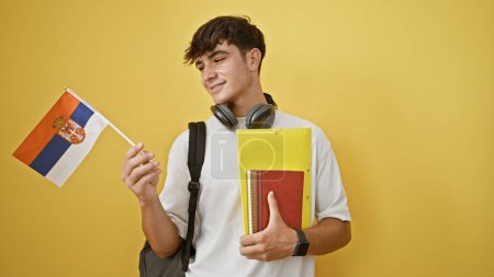 Photo for Confident young hispanic student guy, a proud flag bearer of serbia, passionately holds booking amidst yellow isolated backdrop, embracing education. - Royalty Free Image
