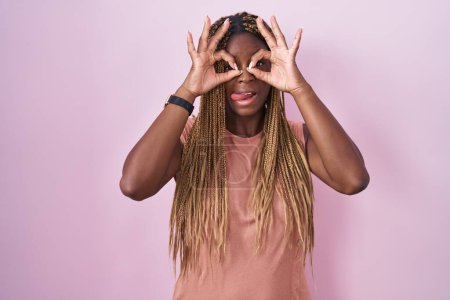 Téléchargez les photos : Femme afro-américaine avec des cheveux tressés debout sur fond rose faisant un geste correct comme des jumelles sortant la langue, les yeux regardant à travers les doigts. expression folle. - en image libre de droit