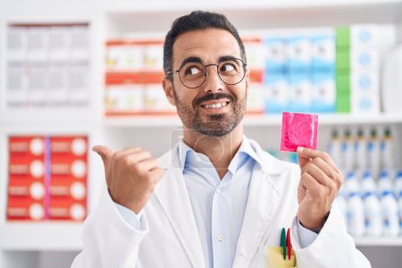 Téléchargez les photos : Homme hispanique avec barbe travaillant à la pharmacie pharmacie tenant préservatif pointant le pouce vers le côté souriant heureux avec la bouche ouverte - en image libre de droit