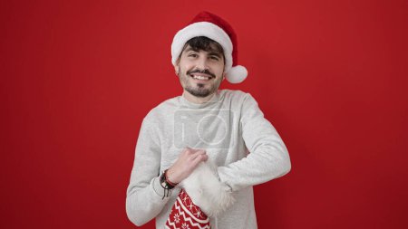 Photo for Young hispanic man wearing christmas hat looking inside of sock over isolated red background - Royalty Free Image