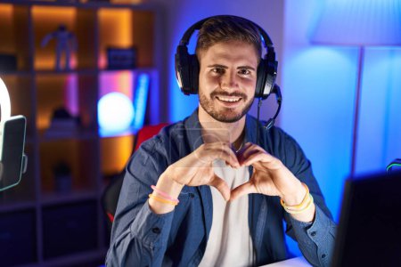 Foto de Young caucasian man streamer smiling confident doing heart symbol with hands at gaming room - Imagen libre de derechos