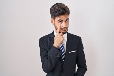Téléchargez les photos : Jeune homme hispanique avec des tatouages portant un costume d'affaires et une cravate pointant vers l'œil vous regardant geste, expression suspecte - en image libre de droit
