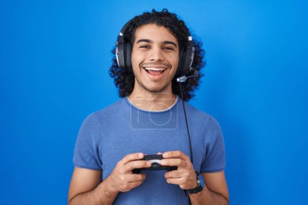Foto de Hombre hispano con pelo rizado jugando videojuego sosteniendo el controlador sonriendo y riendo en voz alta porque divertido chiste loco. - Imagen libre de derechos