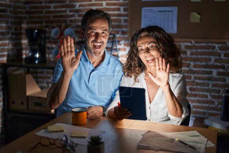Foto de Mediana edad pareja hispana usando touchpad sentado en la mesa por la noche renunciando a decir hola feliz y sonriente, gesto de bienvenida amistoso - Imagen libre de derechos