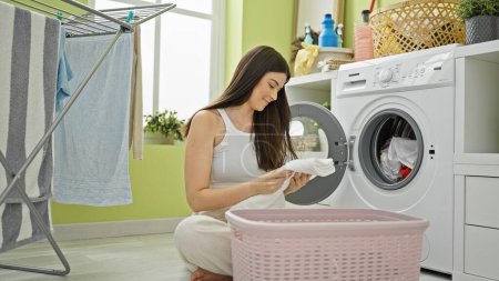 Photo for Young beautiful hispanic woman washing clothes folding towel at laundry room - Royalty Free Image