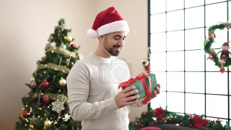 Téléchargez les photos : Jeune homme hispanique tenant cadeau assis sur le canapé près de l'arbre de Noël à la maison - en image libre de droit