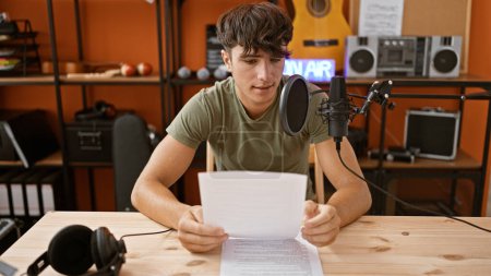 Foto de Adolescente hispano joven guapo dominando el estudio de radio, leyendo y presentando noticias frescas en vivo en el aire con concentración seria, expresión de trabajo - Imagen libre de derechos