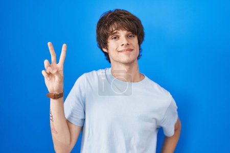 Téléchargez les photos : Jeune homme hispanique debout sur fond bleu souriant en regardant la caméra montrant les doigts faisant signe de victoire. numéro deux. - en image libre de droit