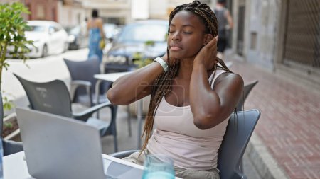 Téléchargez les photos : Femme afro-américaine utilisant ordinateur portable peignage cheveux transpiration à café terrasse - en image libre de droit