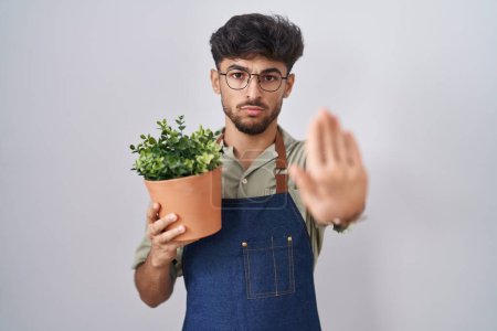 Téléchargez les photos : Homme arabe avec barbe tenant pot de plante verte faire arrêter chanter avec la paume de la main. expression d'avertissement avec geste négatif et sérieux sur le visage. - en image libre de droit