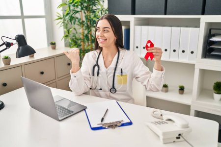 Photo for Hispanic doctor woman holding support red ribbon pointing thumb up to the side smiling happy with open mouth - Royalty Free Image