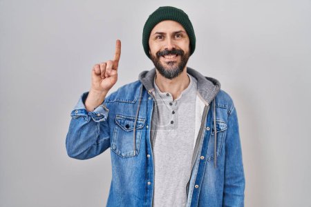 Téléchargez les photos : Jeune homme hispanique avec des tatouages portant une casquette en laine montrant et pointant vers le haut avec le doigt numéro un tout en souriant confiant et heureux. - en image libre de droit