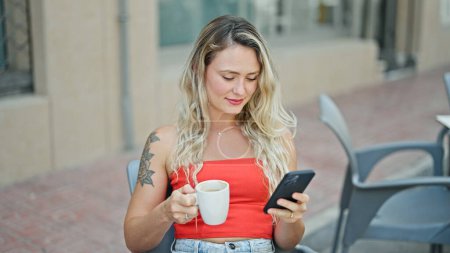 Foto de Mujer rubia joven usando smartphone bebiendo café en la terraza de la cafetería - Imagen libre de derechos