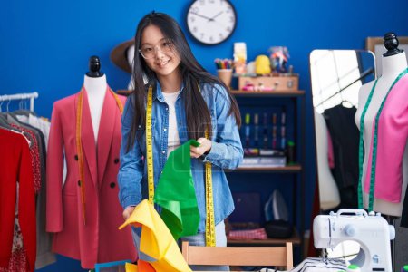 Téléchargez les photos : Jeune femme chinoise tailleur souriant confiance tenant chiffons à l'usine de vêtements - en image libre de droit